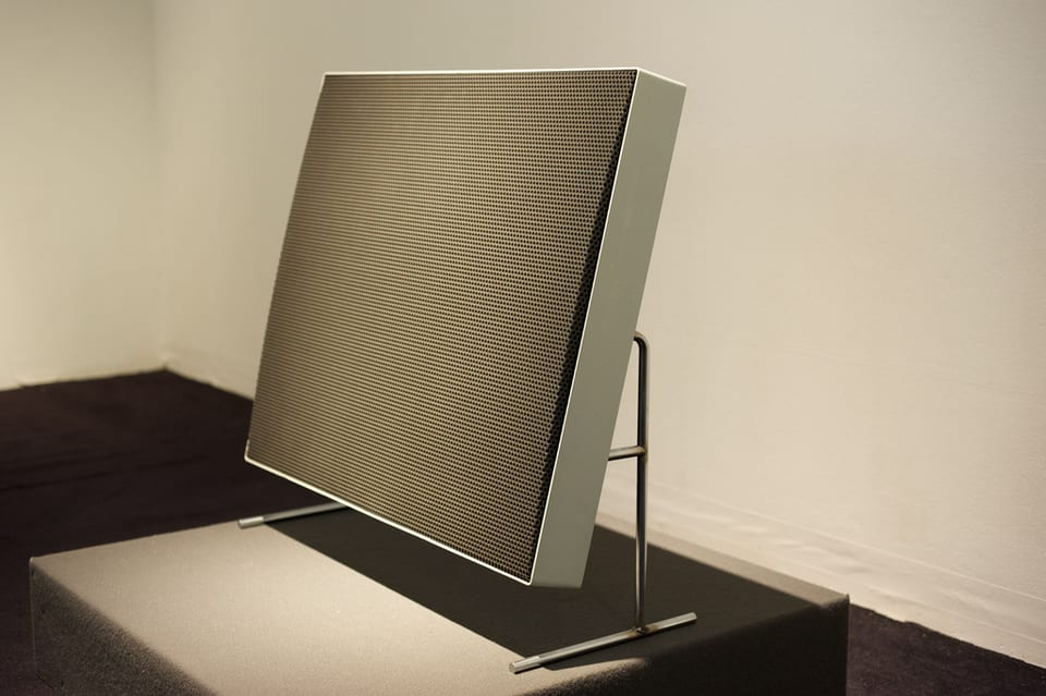 A Braun LE1 flat electrostatic panel loudspeaker on a slim wire stand, its perforated metal stator grille facing the camera.