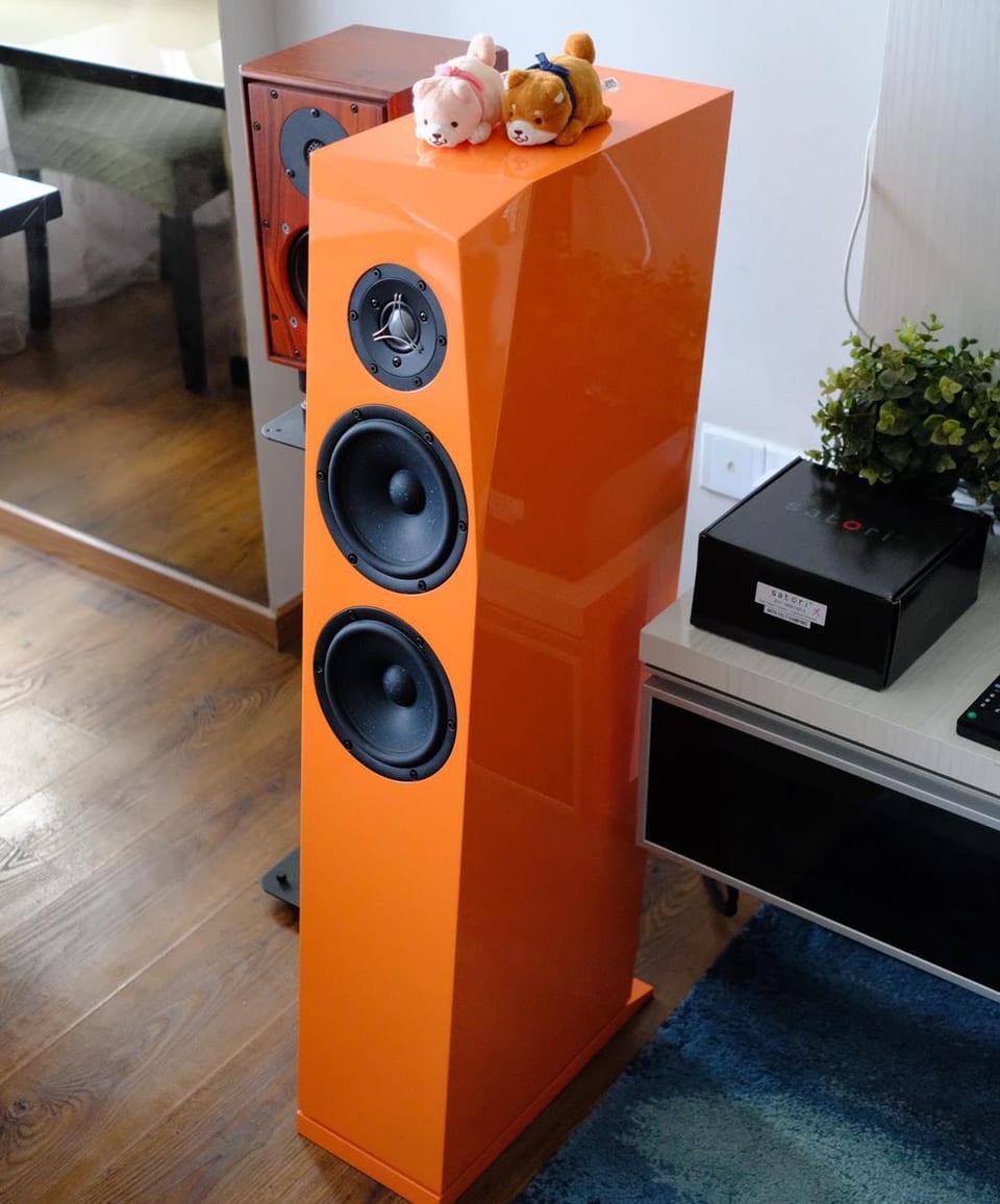 Gloss orange Rinjani Be floorstanding speaker in a living room, one dome tweeter above two black cone drivers