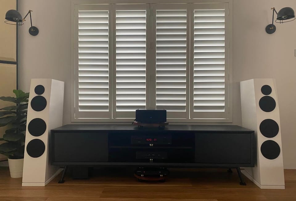 Pair of white SB Acoustics Sasandu floorstanders flanking a dark AV console below shuttered windows