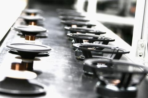 Loudspeaker assembly line: a row of driver chassis and cone assemblies moving down a dark conveyor, each at the same stage of build.