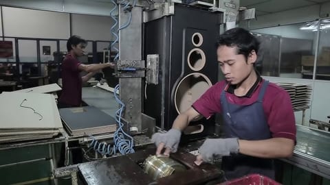 Cabinet work on the factory floor at Sinar Baja Electric: one worker feeding a press in the foreground and another at a machine behind, sheets of MDF stacked to the left, and a finished loudspeaker baffle with three driver cut-outs standing between them.