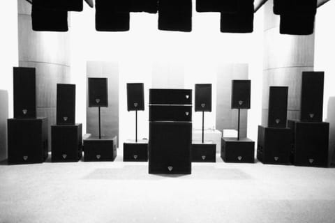 The factory listening room in black and white: banks of professional loudspeaker cabinets and subwoofers arranged along the walls, with absorption panels suspended from the ceiling.